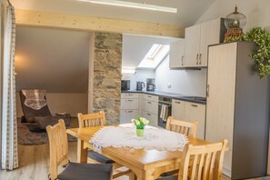 A cozy dining area with a wooden table, chairs, and a lace tablecloth. A stone wall and a kitchen with cabinets and appliances are visible in the background.