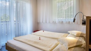 A bedroom with two beds, a window with sheer curtains, a bedside table, and a lamp. - Apartment “Urbanhof - Moritzing” with Mountain View, Wi-Fi & 2 Balconies (Bozen)