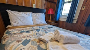 A neatly made bed with a floral bedspread, a red lamp on a wooden nightstand, and a window with blue curtains. - Romatic Retreat at the Honeymoon Sunday Cottage located at the Oceana Cottages (North Truro)