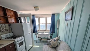 A compact room with a kitchenette, a bed with floral pillows, a window with curtains, and a view of the sea. - Romatic Retreat at the Honeymoon Sunday Cottage located at the Oceana Cottages (North Truro)