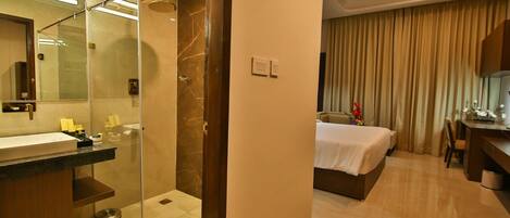 A modern hotel room with a glass-enclosed shower, a large mirror, a bed with a white bedspread, a desk with a chair, and a small trash bin.