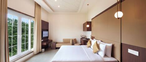 A hotel room with a large bed, a TV, a desk, and a view of greenery through the window.