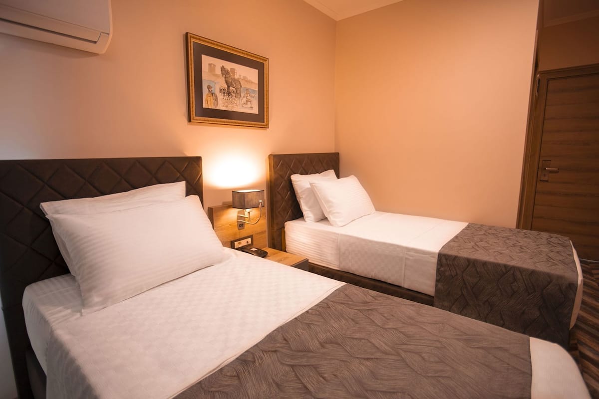Habitación de hotel con dos camas, una mesita de noche con lámpara, un cuadro en la pared y una puerta de madera.