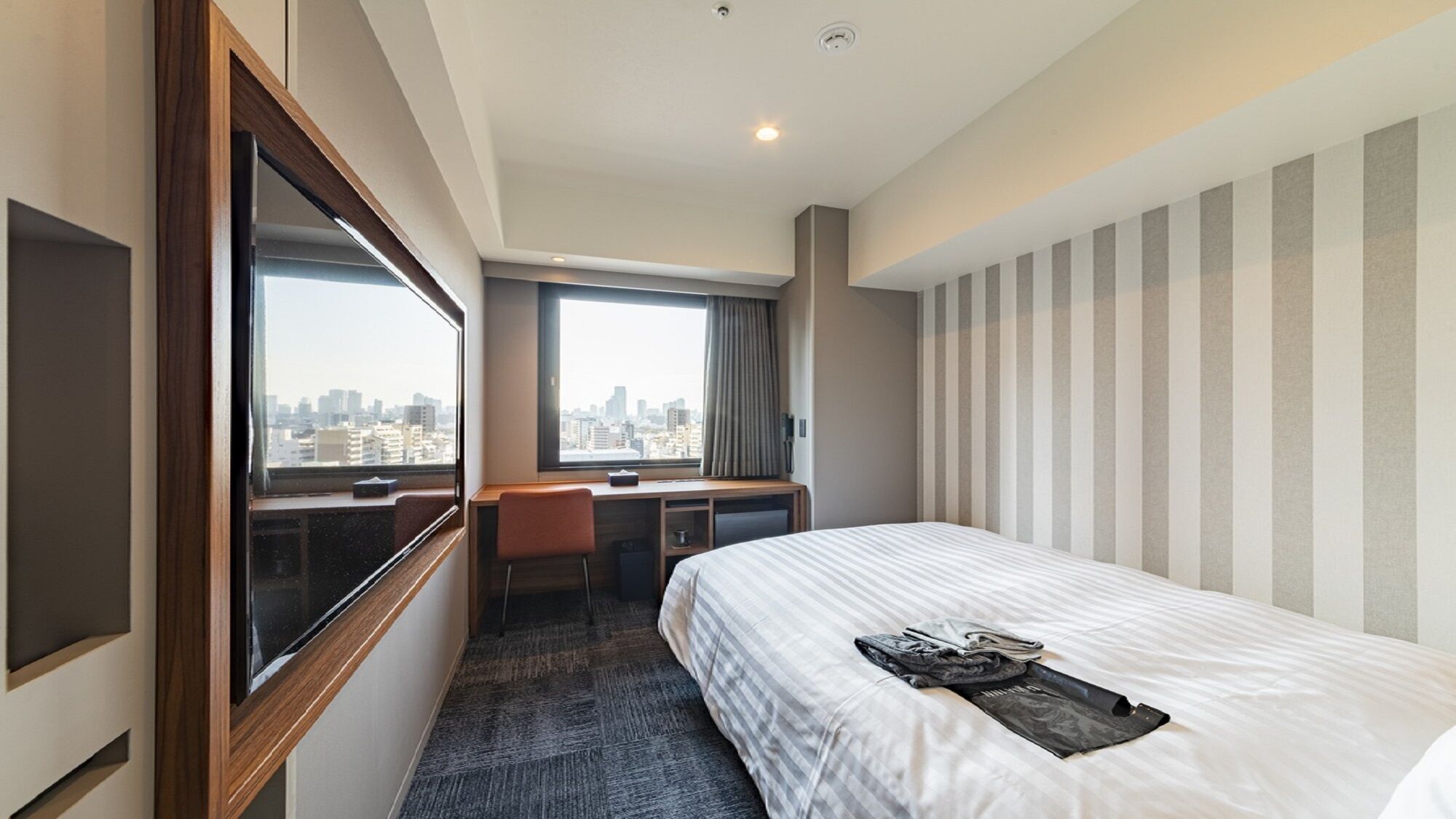 A hotel room with a bed, a desk, a chair, a window with a city view, and a striped wall.