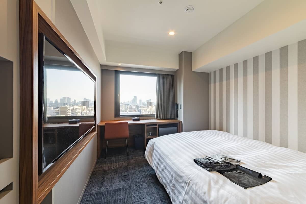 A hotel room with a bed, a desk, a chair, a window with a city view, and a striped wall.