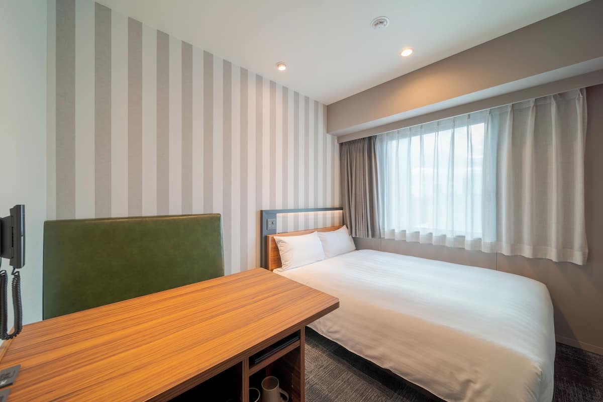 A hotel room with a single bed, a wooden desk, a green upholstered chair, a flat-screen TV, and a window with curtains.