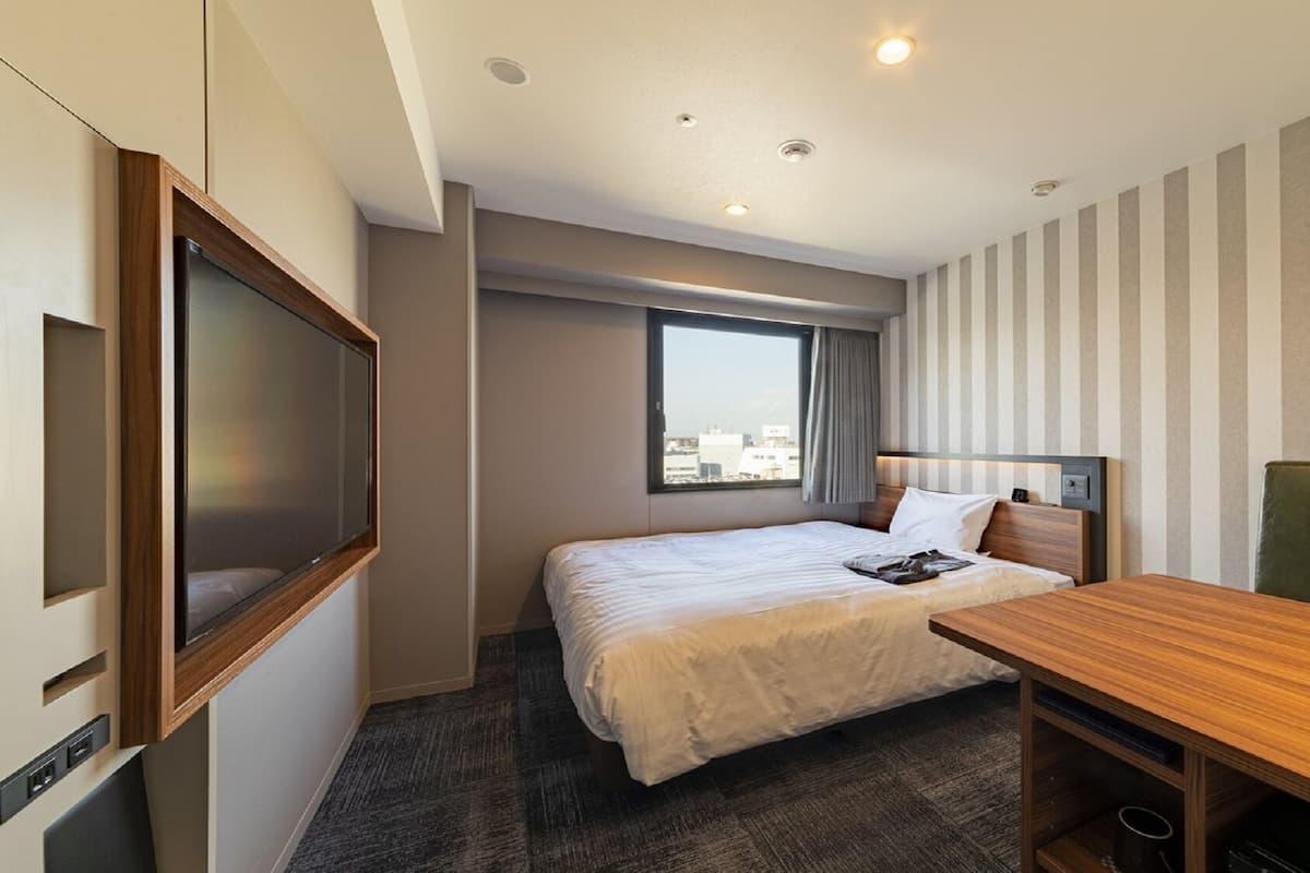 A hotel room with a bed, a desk, a television, and a window with a city view.