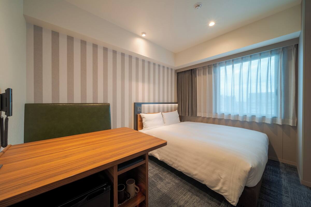 A hotel room with a bed, a desk, a chair, a TV, and a window with curtains.