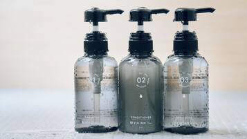 Três garrafas transparentes com bombas dosadoras, identificadas com os números 01, 02 e 03, contendo shampoo, condicionador e sabonete líquido, respectivamente.