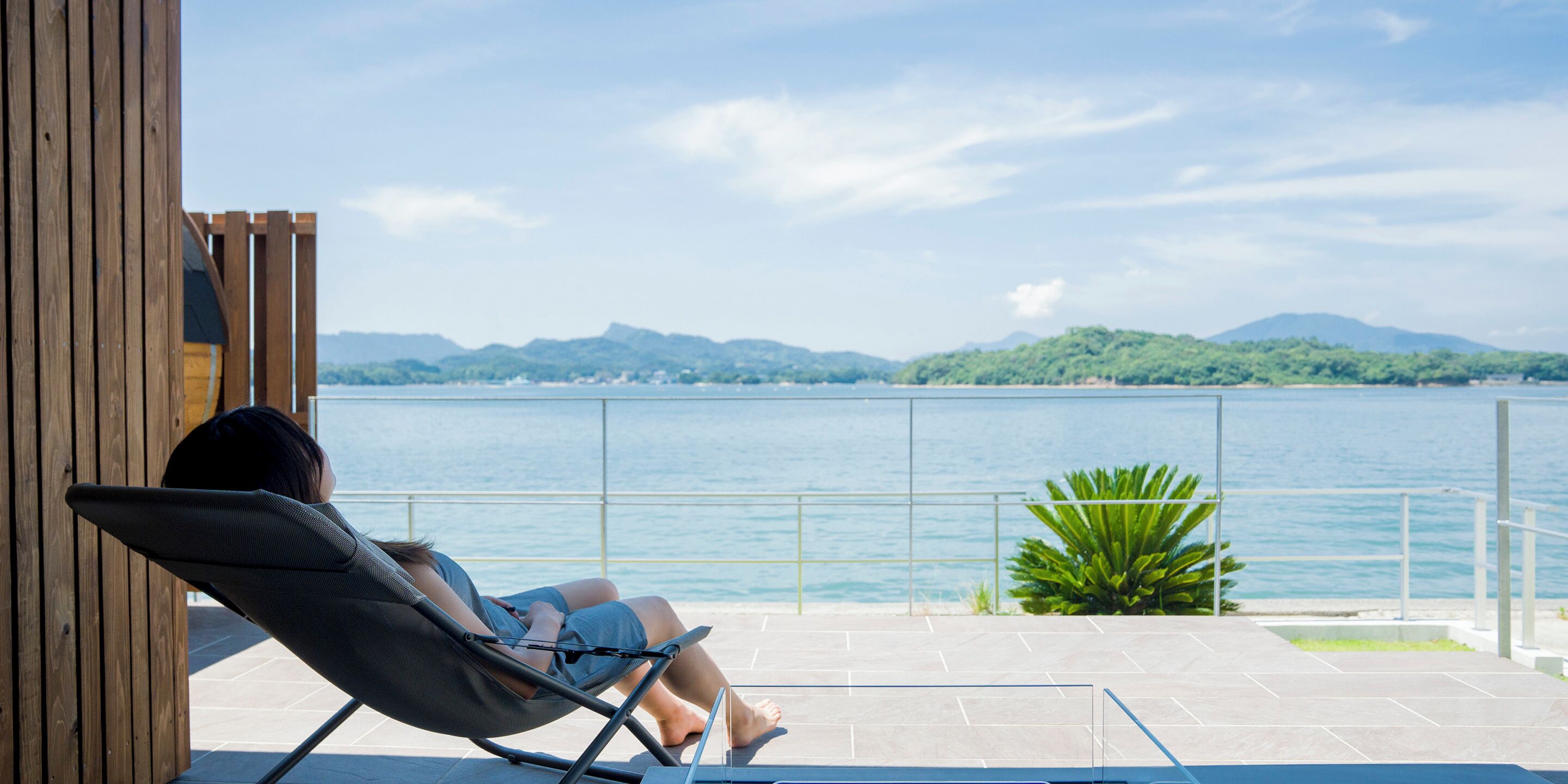 Uma pessoa relaxando em uma espreguiçadeira à beira da piscina, com vista para um lago e montanhas ao longe.