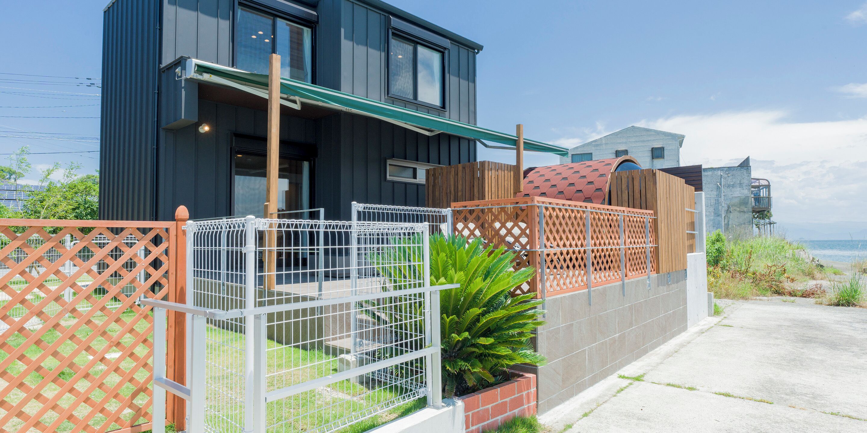 A modern two-story house with a balcony, a fenced garden area, and a view of the sea.