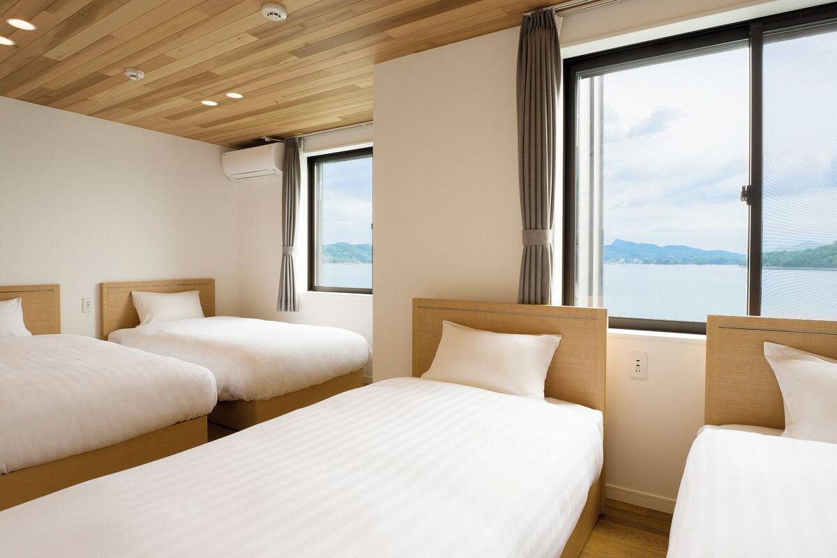 Une chambre d’hôtel avec trois lits, un plafond en bois, de grandes fenêtres donnant sur un lac et des montagnes, et un appareil de climatisation.