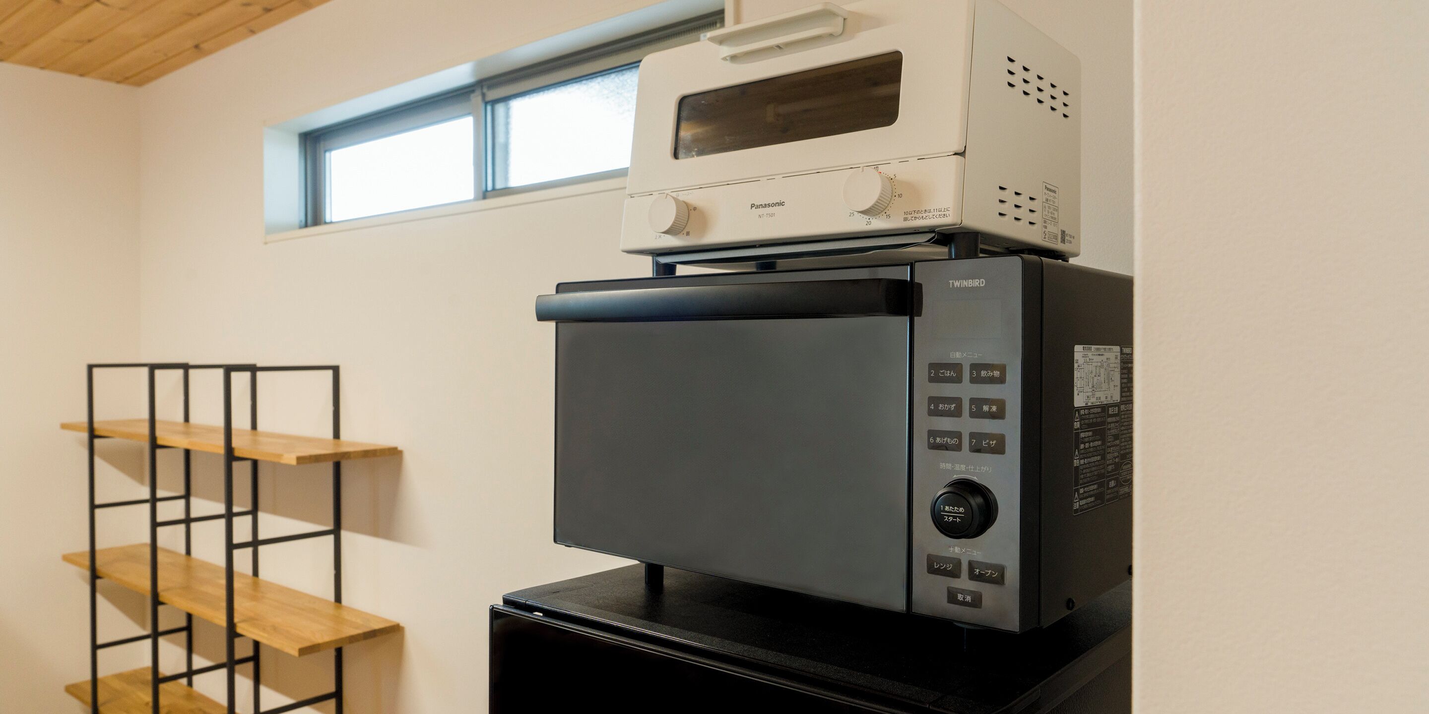A kitchen with a microwave, toaster, and refrigerator. There is a window above the appliances and a wooden shelf to the left.