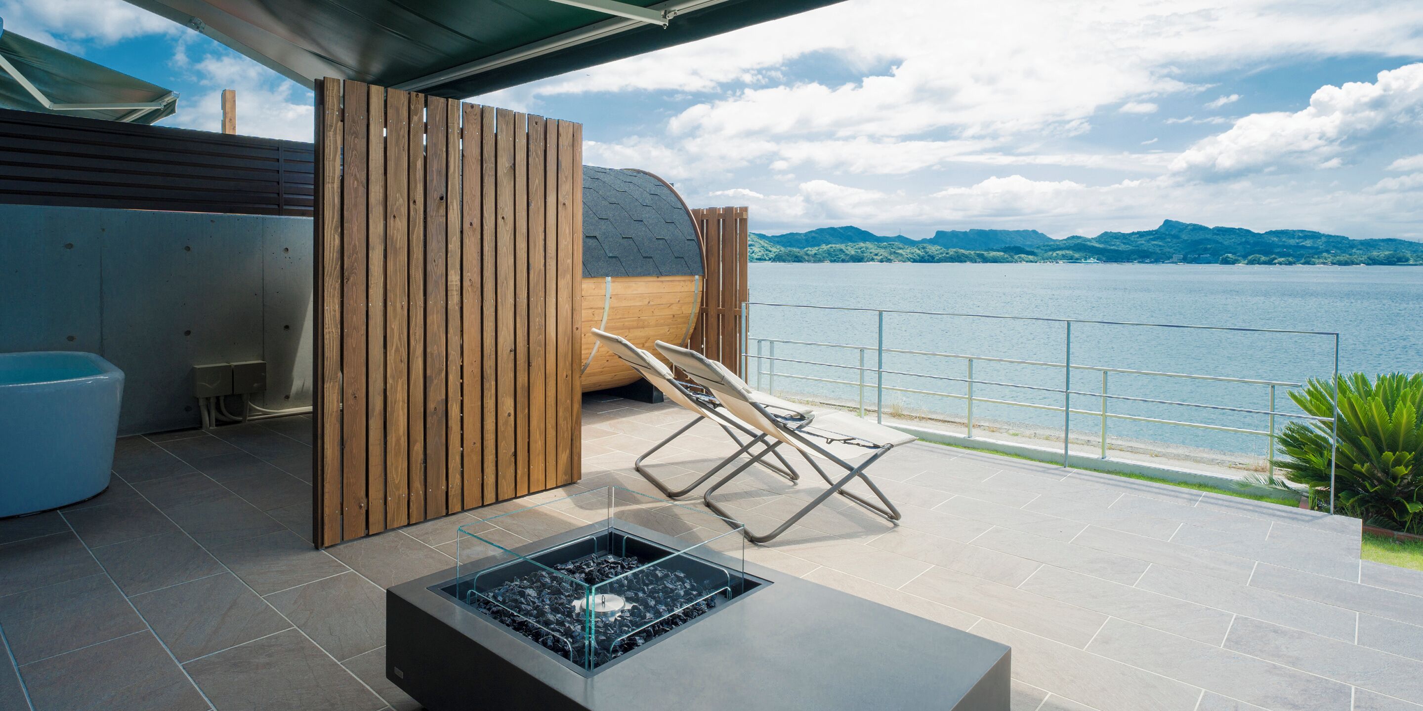 Ein moderner Außenbereich mit Sitzgelegenheit, einem Feuerkorb, einer Holz-Privatsphäre und Blick auf einen See und Berge.