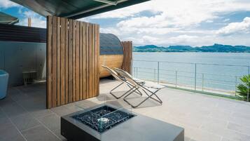 Ein moderner Außenbereich mit Sitzgelegenheit, einem Feuerkorb, einer Holz-Privatsphäre und Blick auf einen See und Berge.