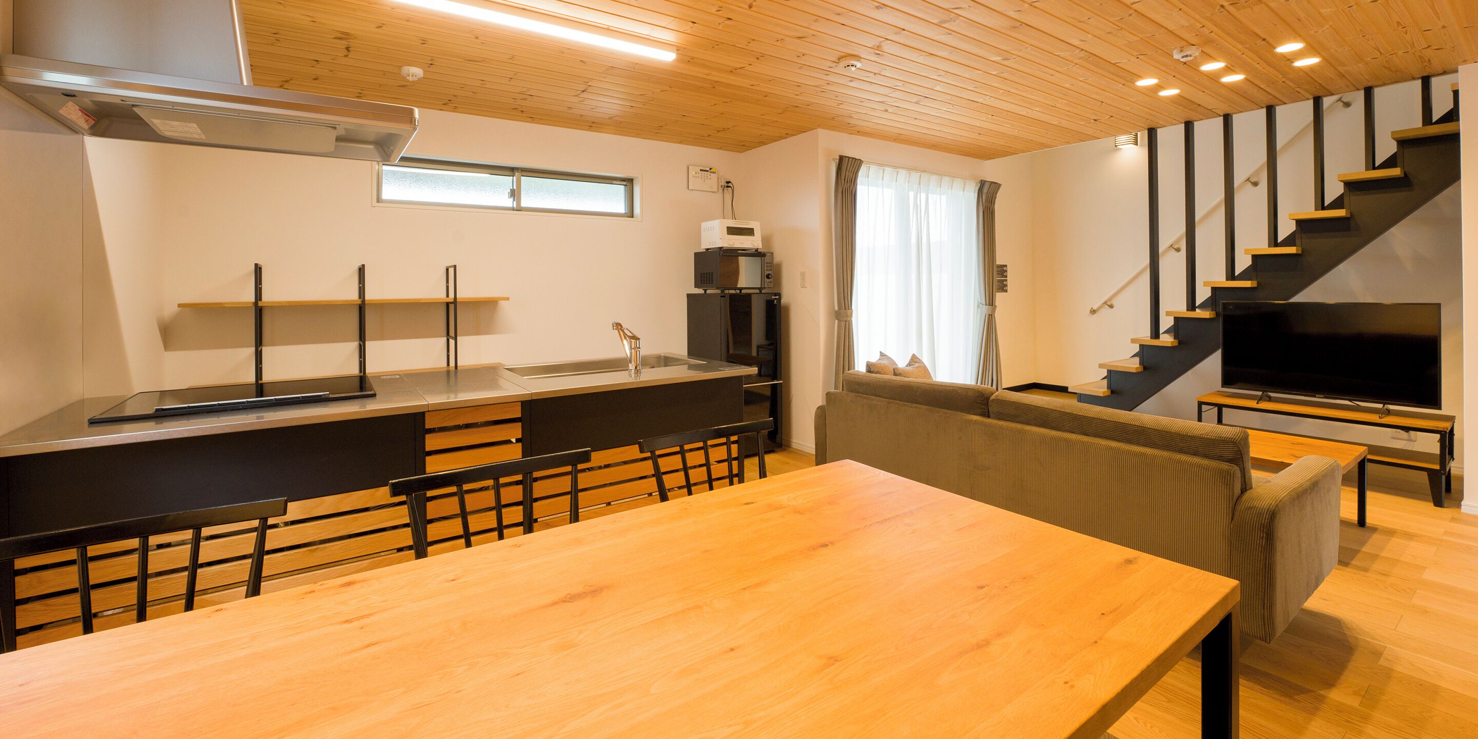A modern kitchen with a wooden table and chairs, a built-in oven, and a TV in the living area.