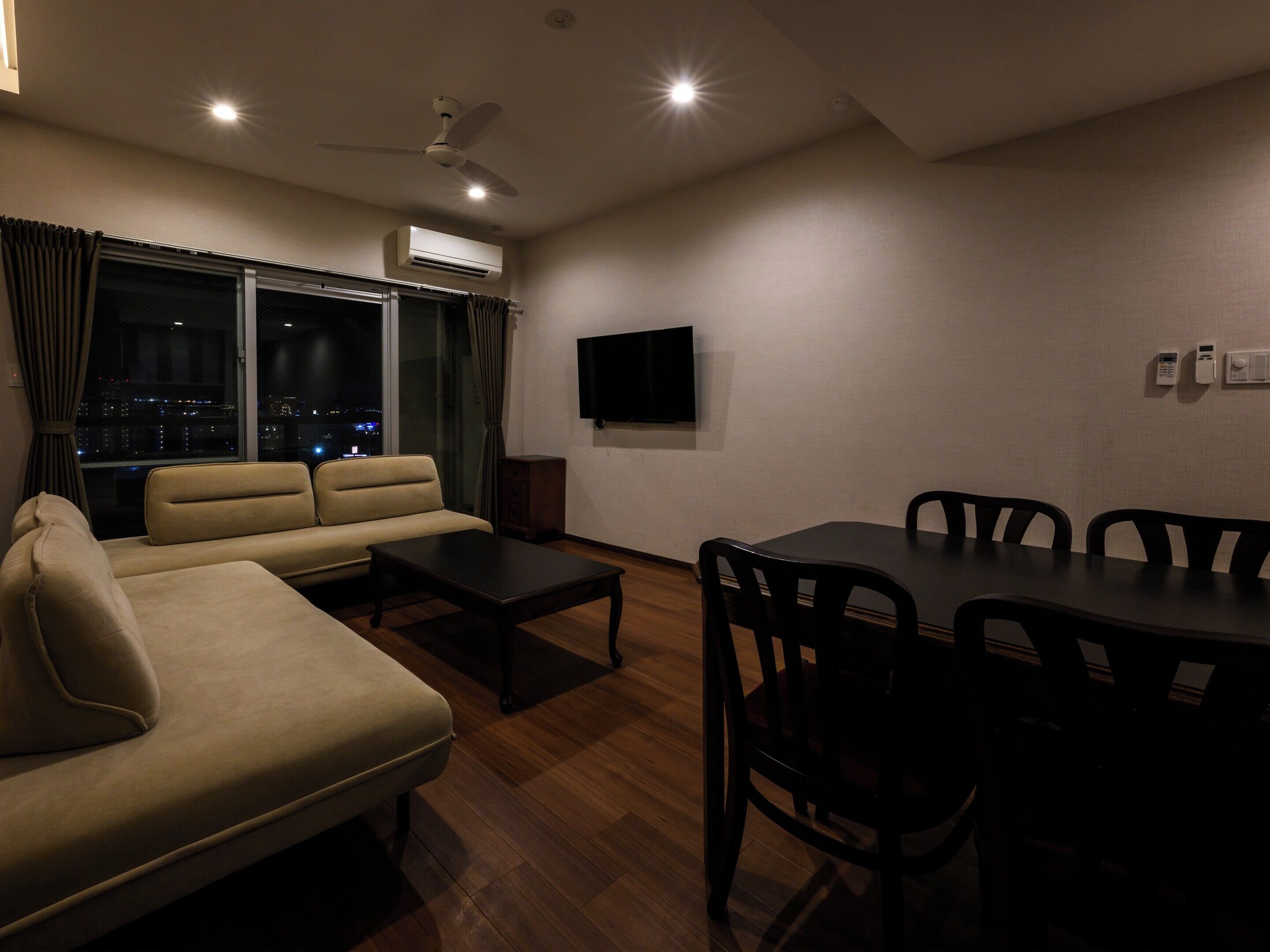 A modern living room with a sofa, a coffee table, a dining table with chairs, a flat-screen TV, and an air conditioning unit.