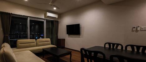 A modern living room with a sofa, a coffee table, a dining table with chairs, a flat-screen TV, and an air conditioning unit.