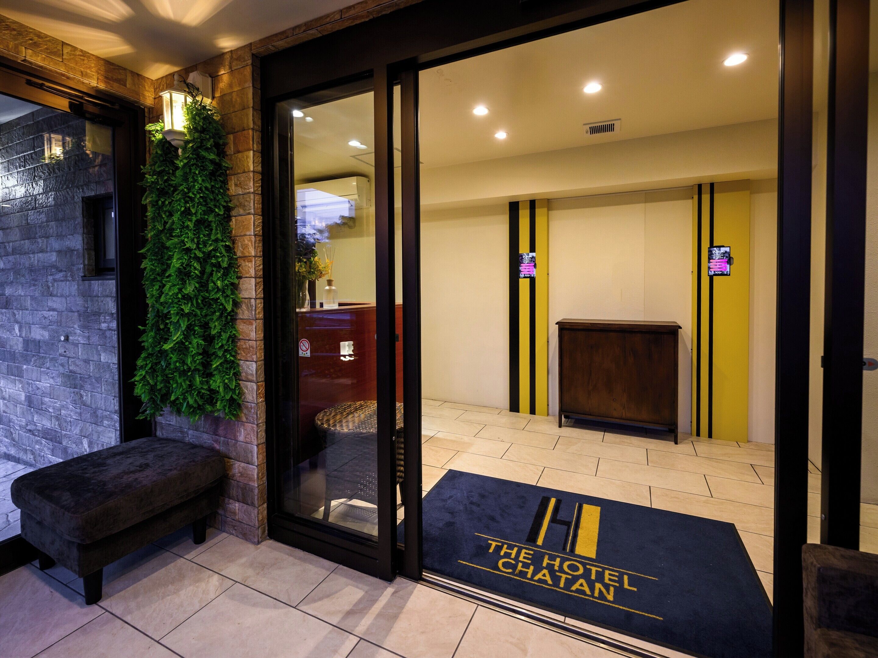 A hotel entrance with a bench, a glass door, a reception desk, and a doormat with the hotel name.