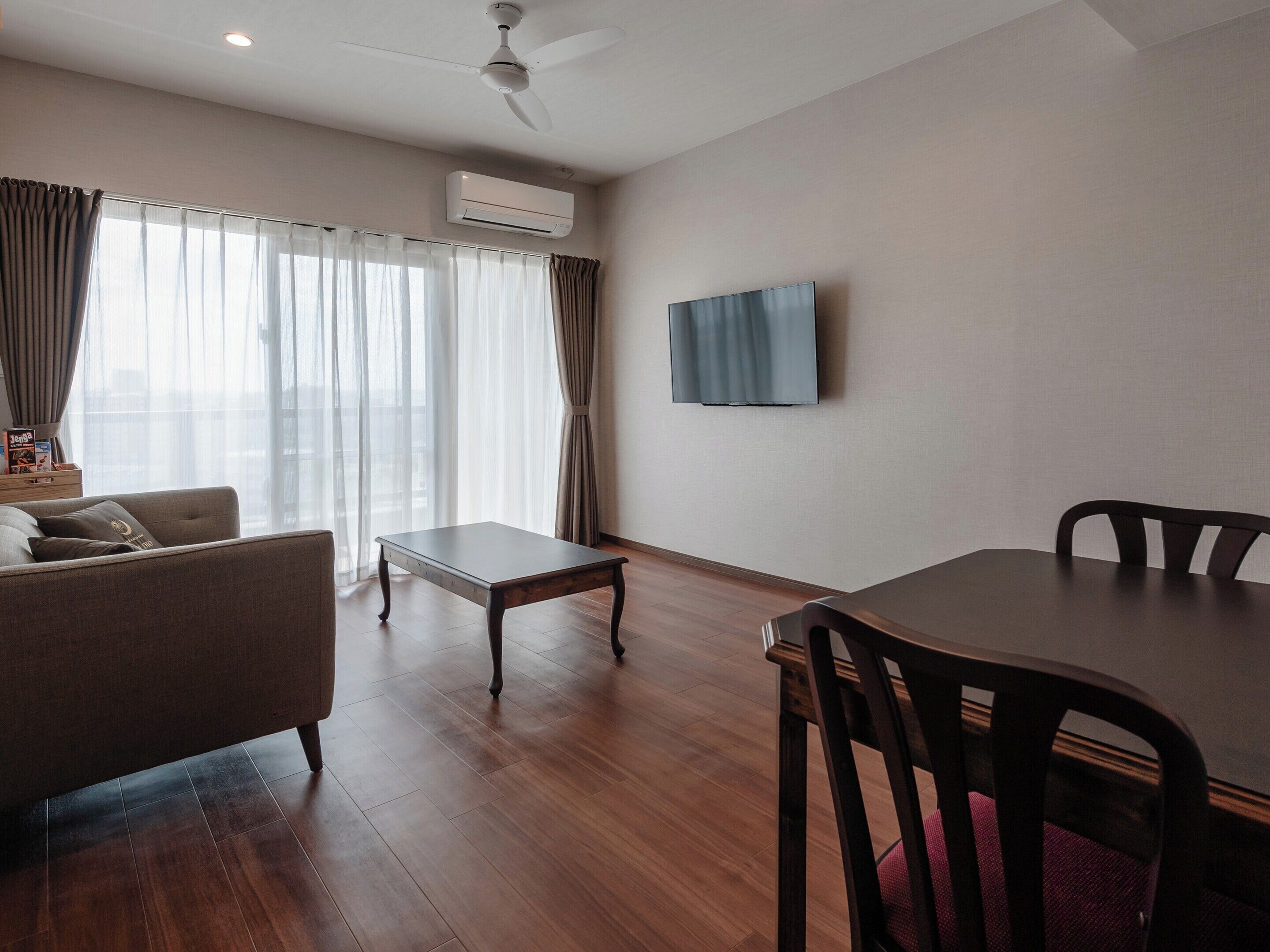 A modern living room with a sofa, a coffee table, a dining table with chairs, a flat-screen TV mounted on the wall, and an air conditioning unit.