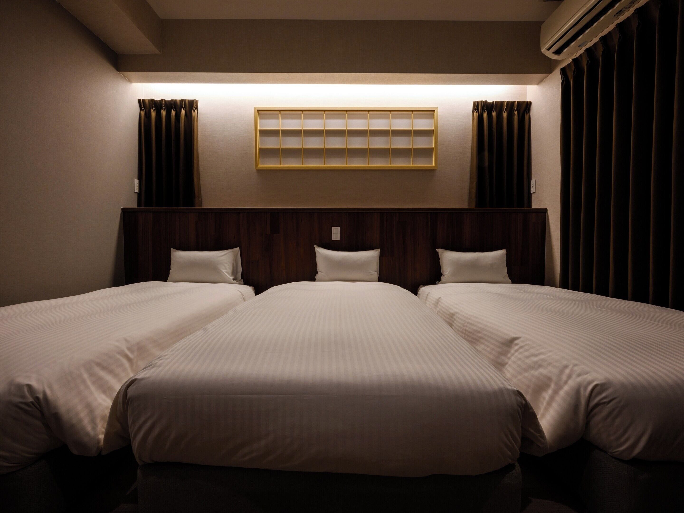 A hotel room with three beds, a headboard, a wall-mounted shelf, and curtains.