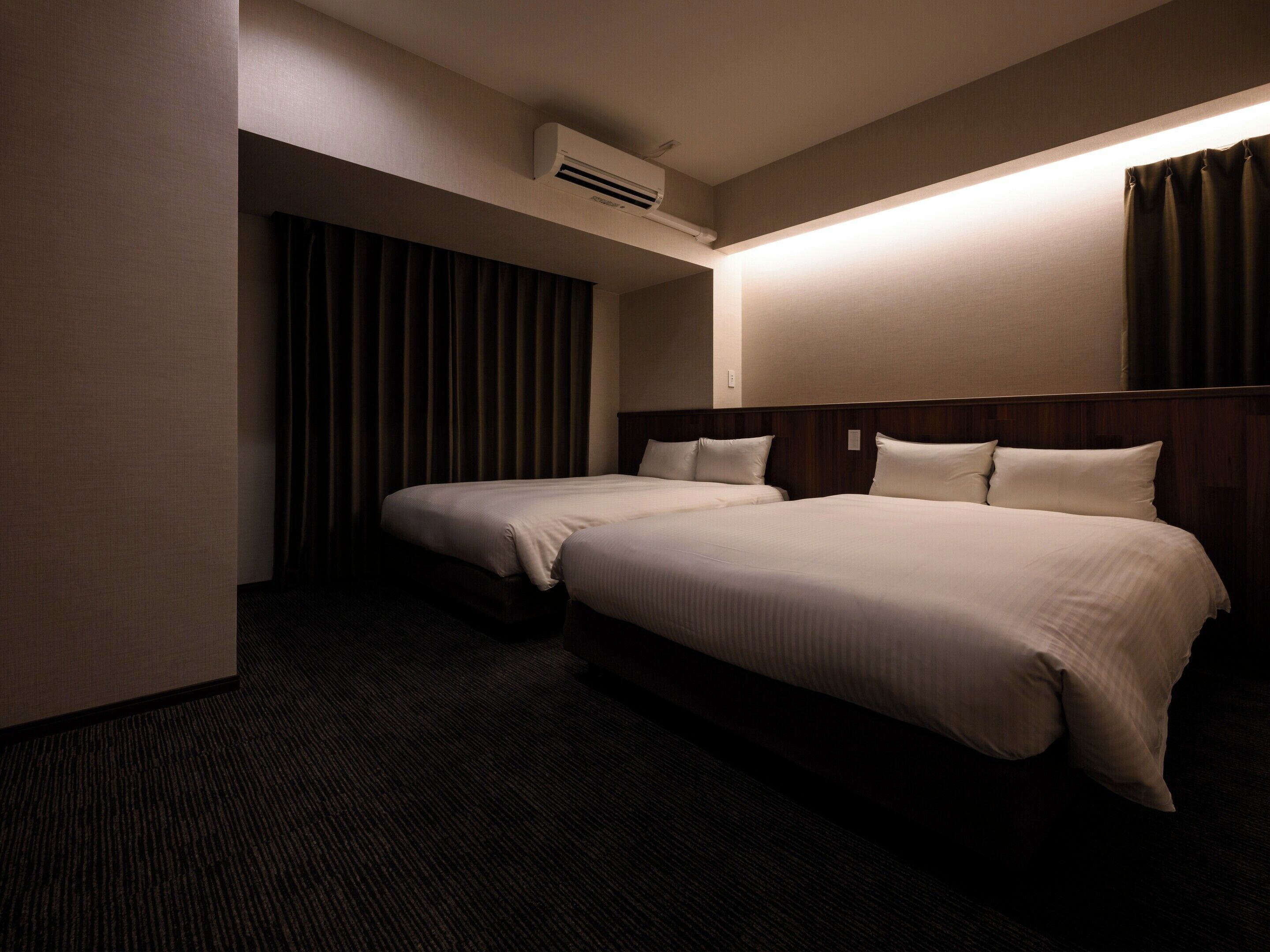 A hotel room with two beds, a dark carpet, and an air conditioning unit on the wall.