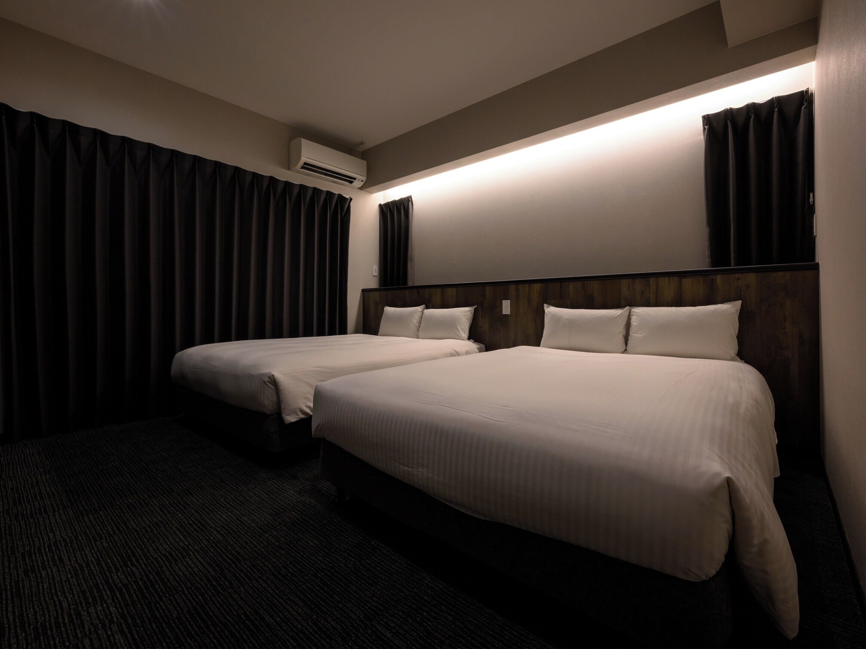 A hotel room with two beds, dark curtains, an air conditioning unit, and a wall-mounted light.