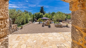 Un espace extérieur aménagé en terrasse, doté d’un sol en pierre, entouré de verdure et agrémenté d’une pergola surmontée d’un parasol violet.