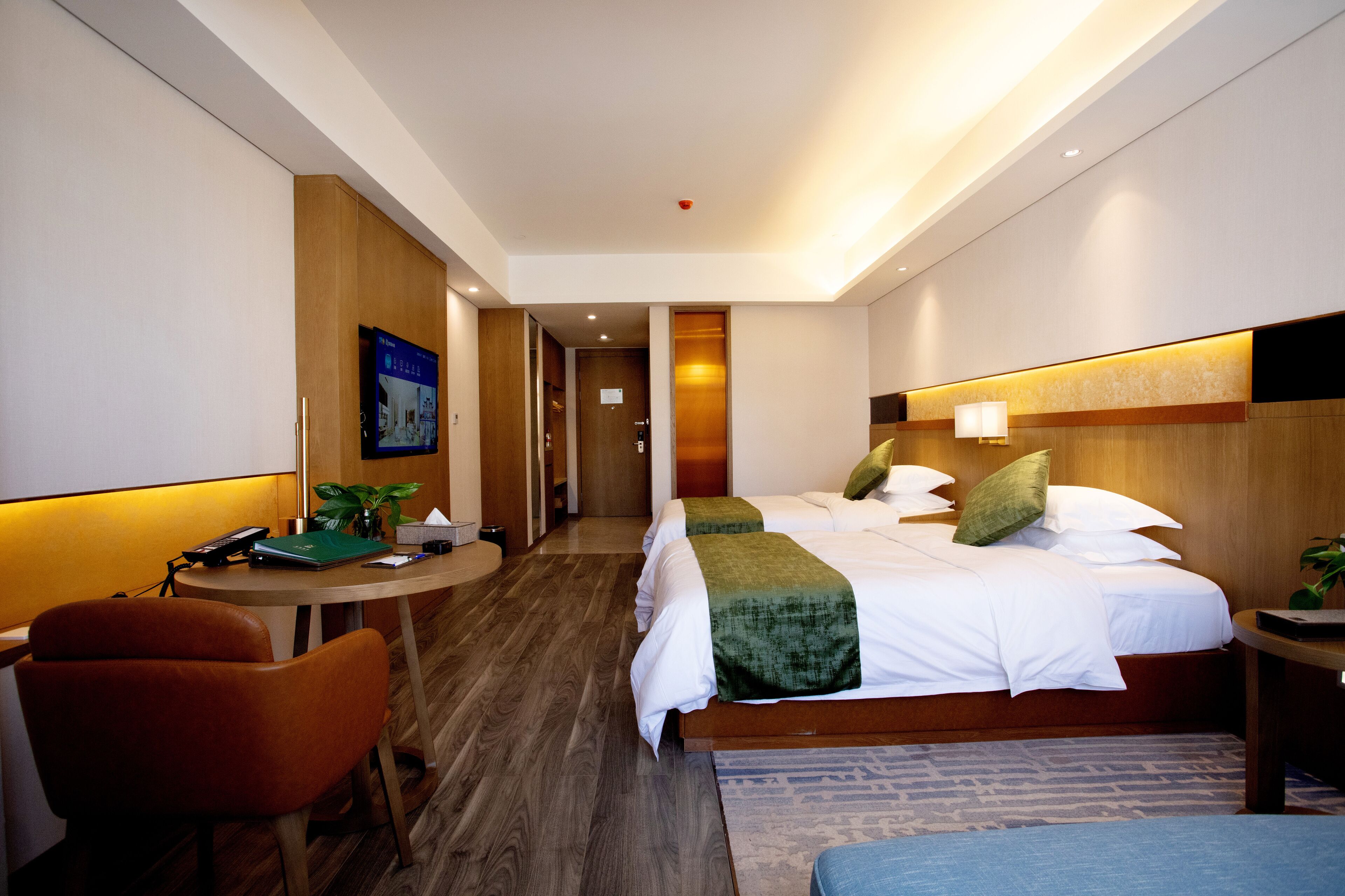 A hotel room with a desk, two beds, a chair, and a small table with a plant.