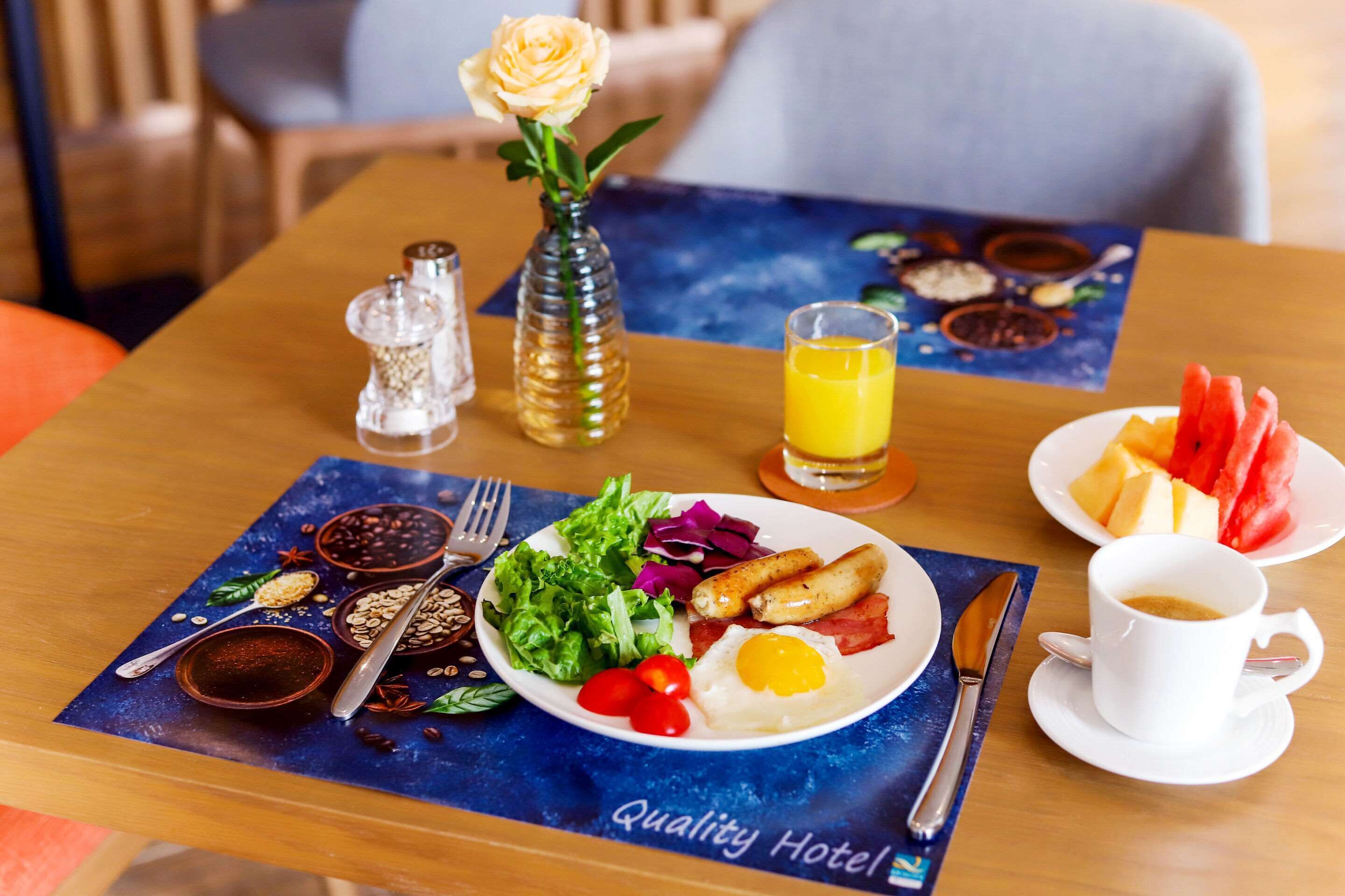 Een ontbijttafel met een bord eieren, spek, tomaten en groenten, een glas sinaasappelsap en een kop koffie, alles op een tafel met een blauwe placemat.