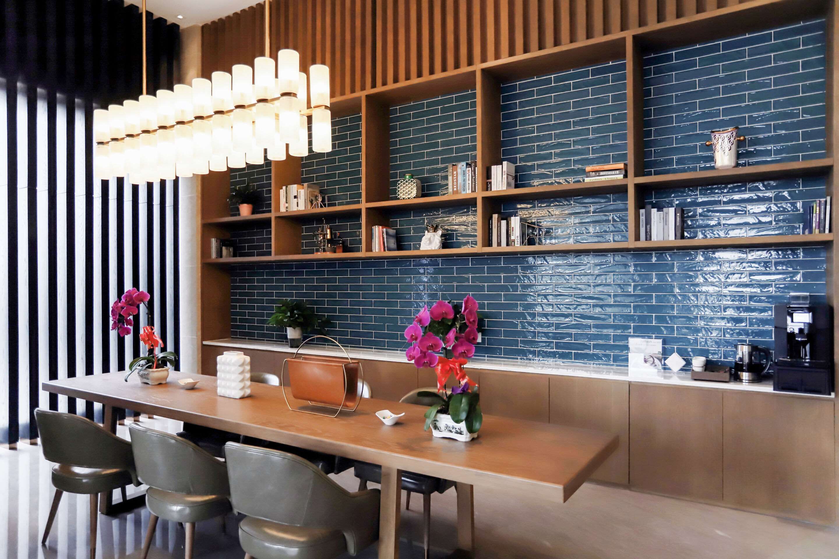 A modern dining area with a wooden table, grey chairs, a hanging light fixture, and a brick-patterned wall with shelves.