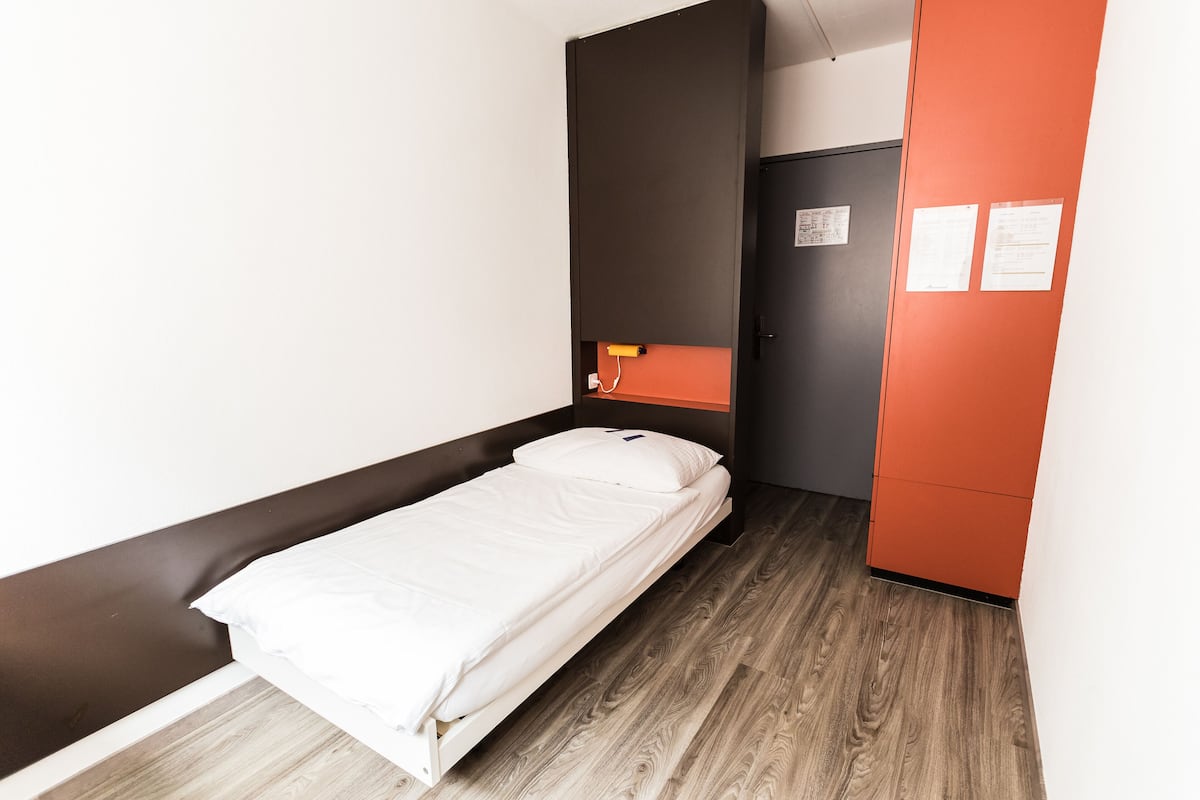 A single bed in a room with a wooden floor, a black wall, and an orange cabinet.