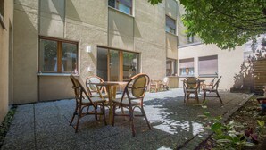 Un espace extérieur aménagé avec des tables et des chaises, un bâtiment doté de grandes fenêtres et un jardin avec des plantes.