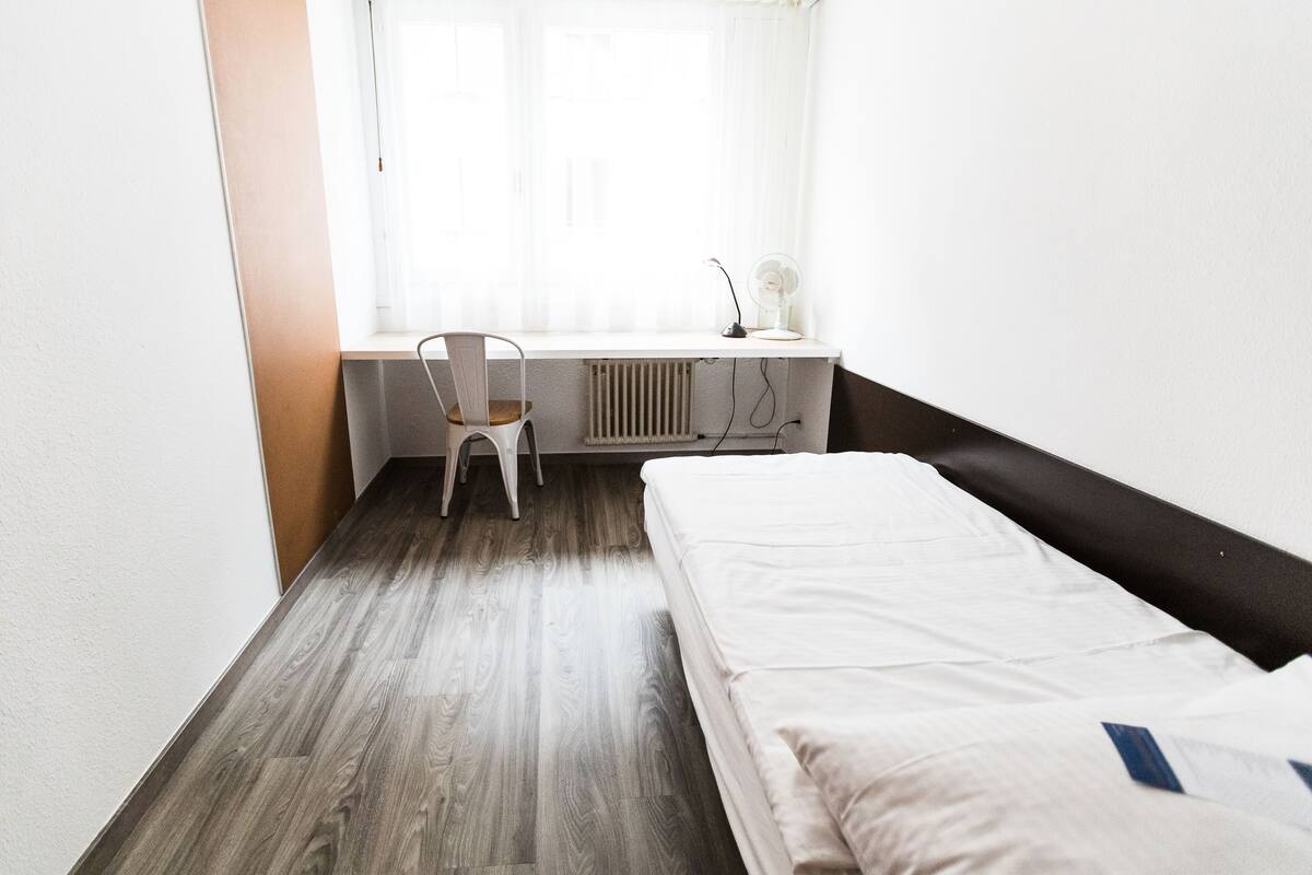 A small, clean room with a single bed, a chair, a desk with a lamp, and a radiator.