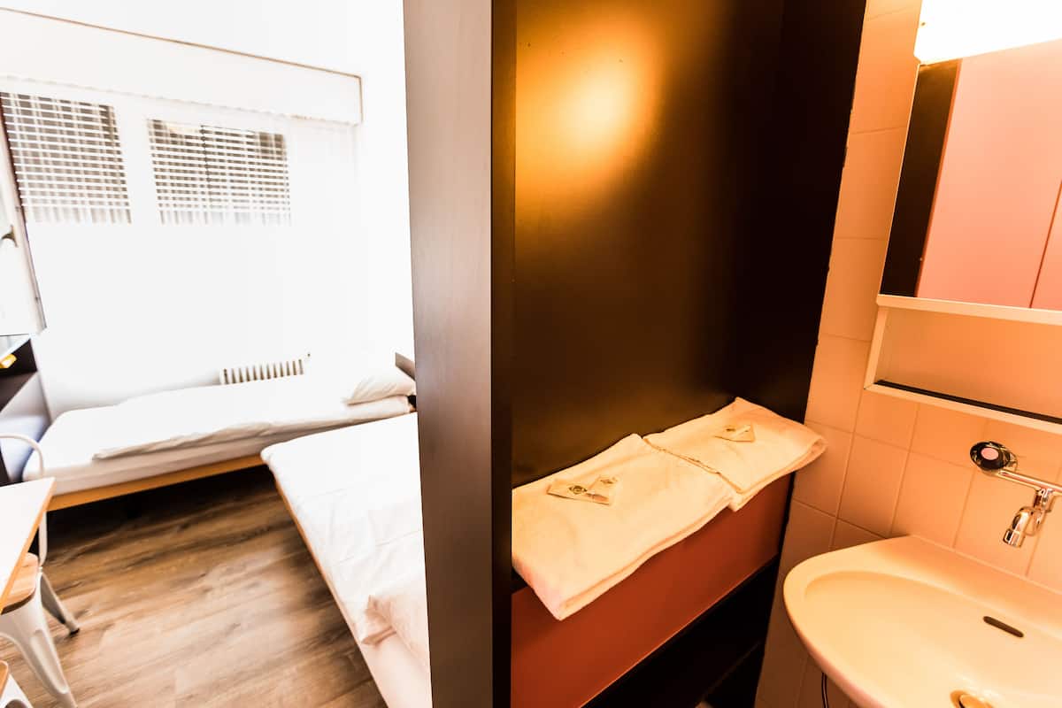 A hotel room with a bed, a bedside table, a small desk, a chair, a window with blinds, a bathroom with a sink and mirror, and a towel hanging on the wall.