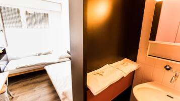 Habitación de hotel con cama, mesita de noche, escritorio pequeño, silla, ventana con persianas, baño con lavabo y espejo, y una toalla colgada en la pared.