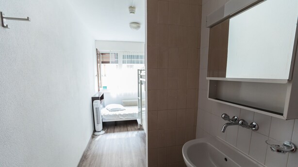A narrow hallway leading to a bedroom with a white bed and a window with blinds. A bathroom with a sink and mirror is visible to the right. - City Hostel Geneva (Geneva)