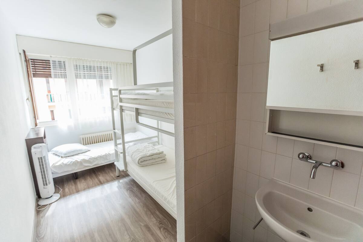 A room with bunk beds, a white radiator, a white sink, and a window with blinds.