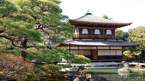 Un edificio japonés tradicional con techo curvo, rodeado por un estanque con rocas y árboles de pino.