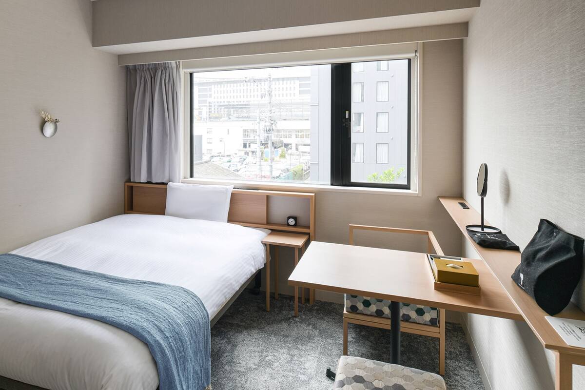 Habitación de hotel con cama, escritorio, silla, mesita, espejo y ventana con vistas a edificios.