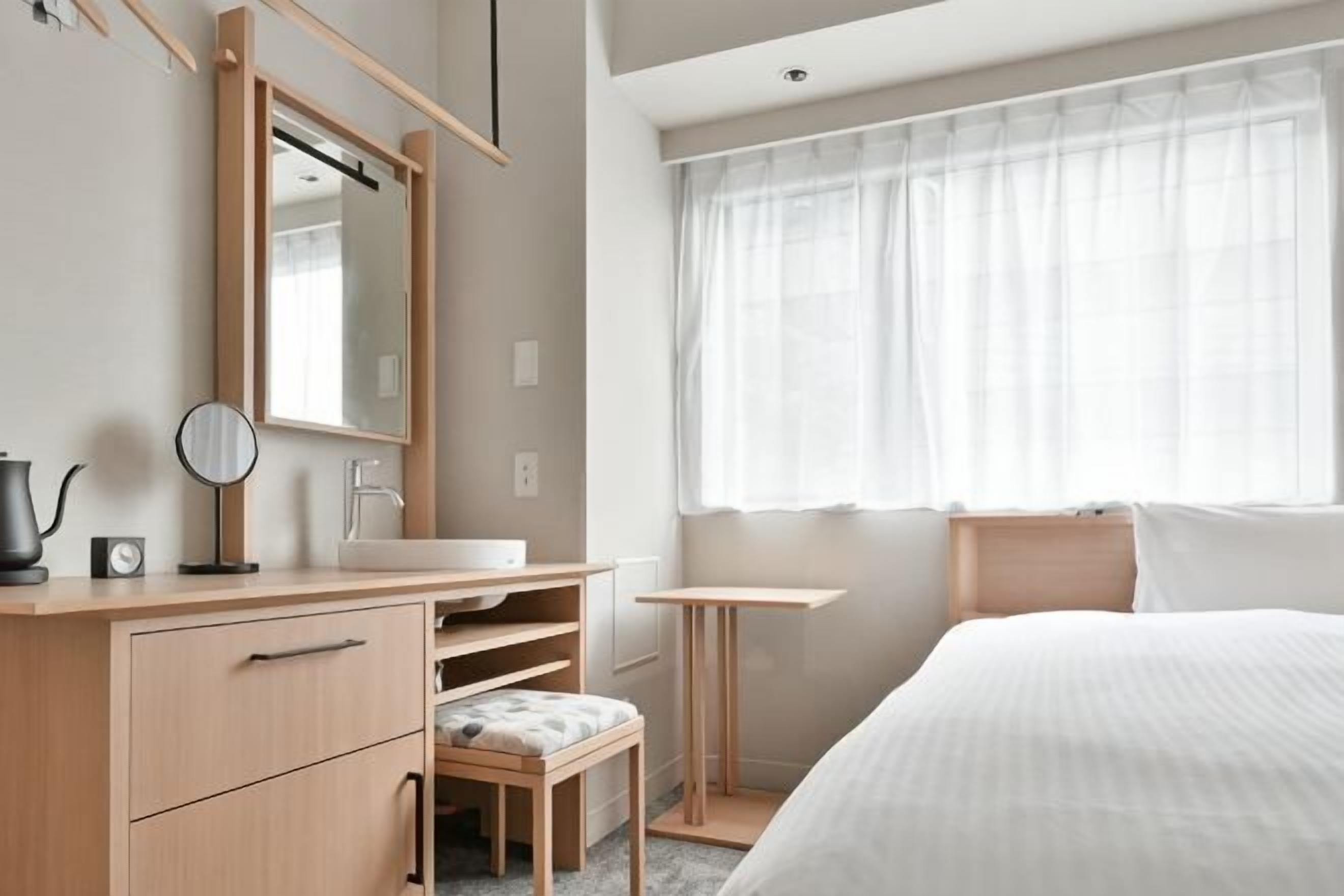 Une chambre à coucher comprenant un lit, une table de chevet, un miroir, une commode et une fenêtre avec des rideaux légers.