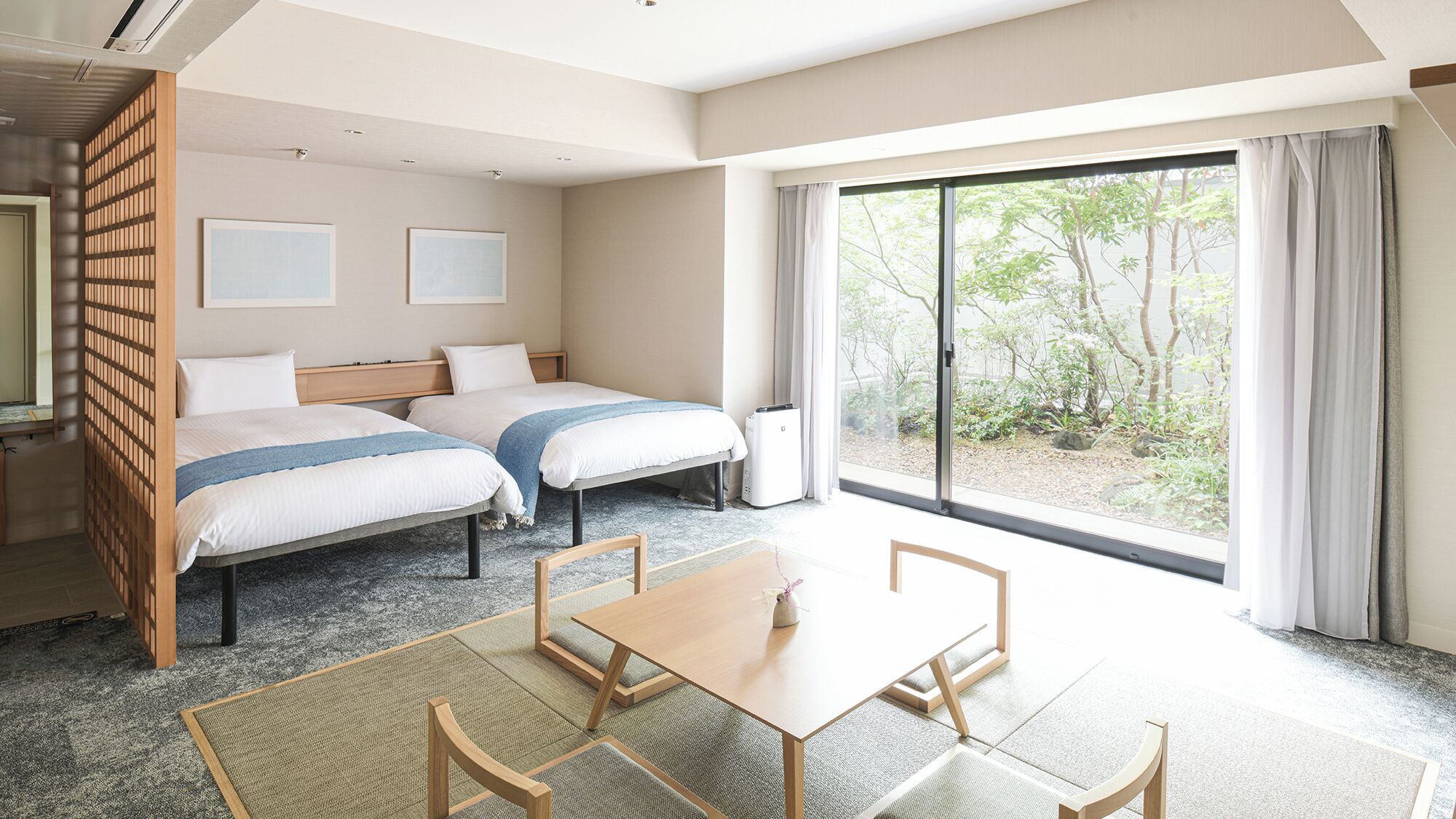 Habitación de hotel con dos camas, una mesa de comedor, sillas, un ventanal corredero y vistas a zonas verdes.