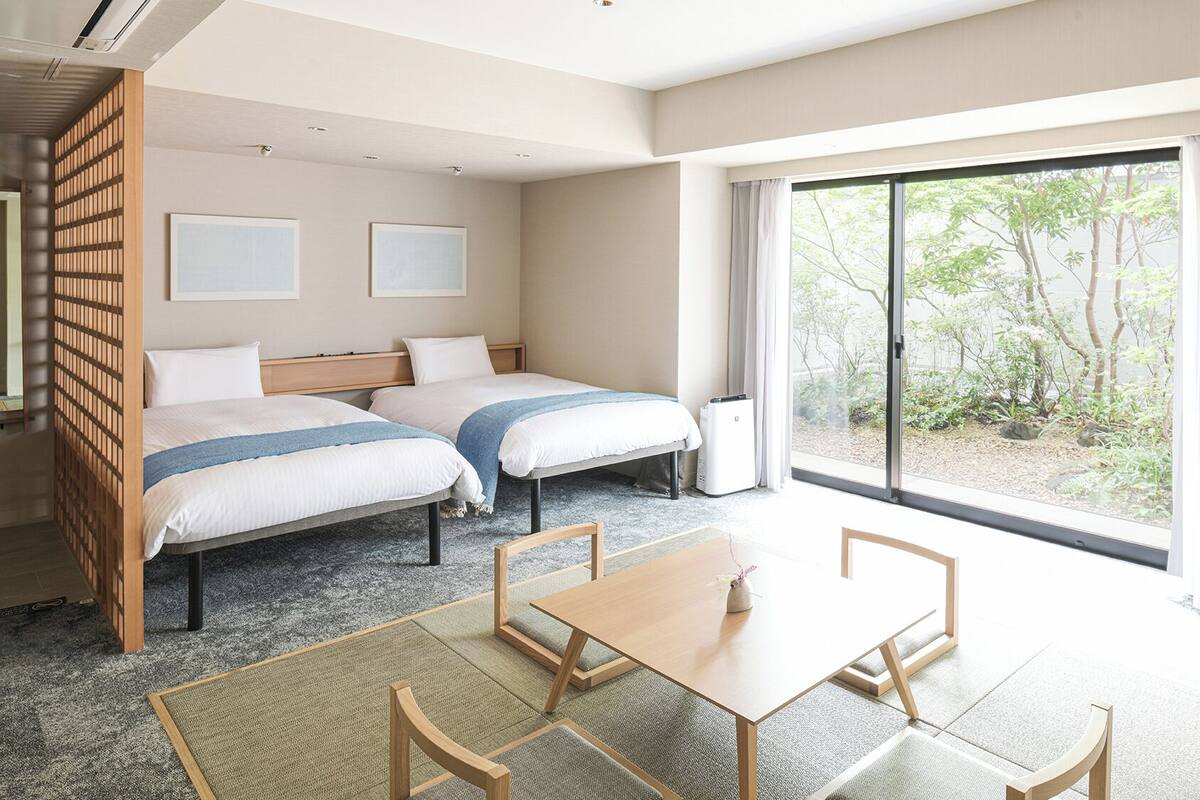 Habitación de hotel con dos camas, una mesa de comedor, sillas, un ventanal corredero y vistas a zonas verdes.