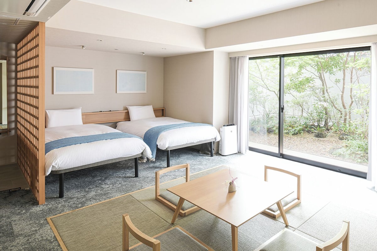A hotel room with two beds, a dining table, chairs, a sliding glass door, and a view of greenery outside.
