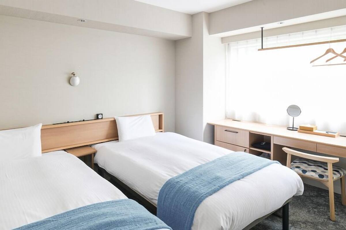 Habitación de hotel con dos camas, un escritorio, una silla, una lámpara colgante y ventana con vistas.