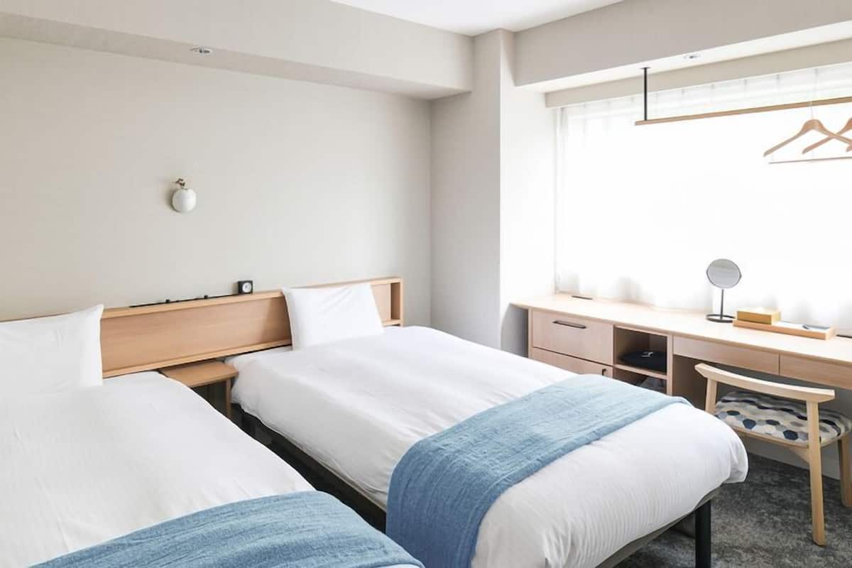 A hotel room with two beds, a desk, a chair, a hanging light fixture, and a window with a view.