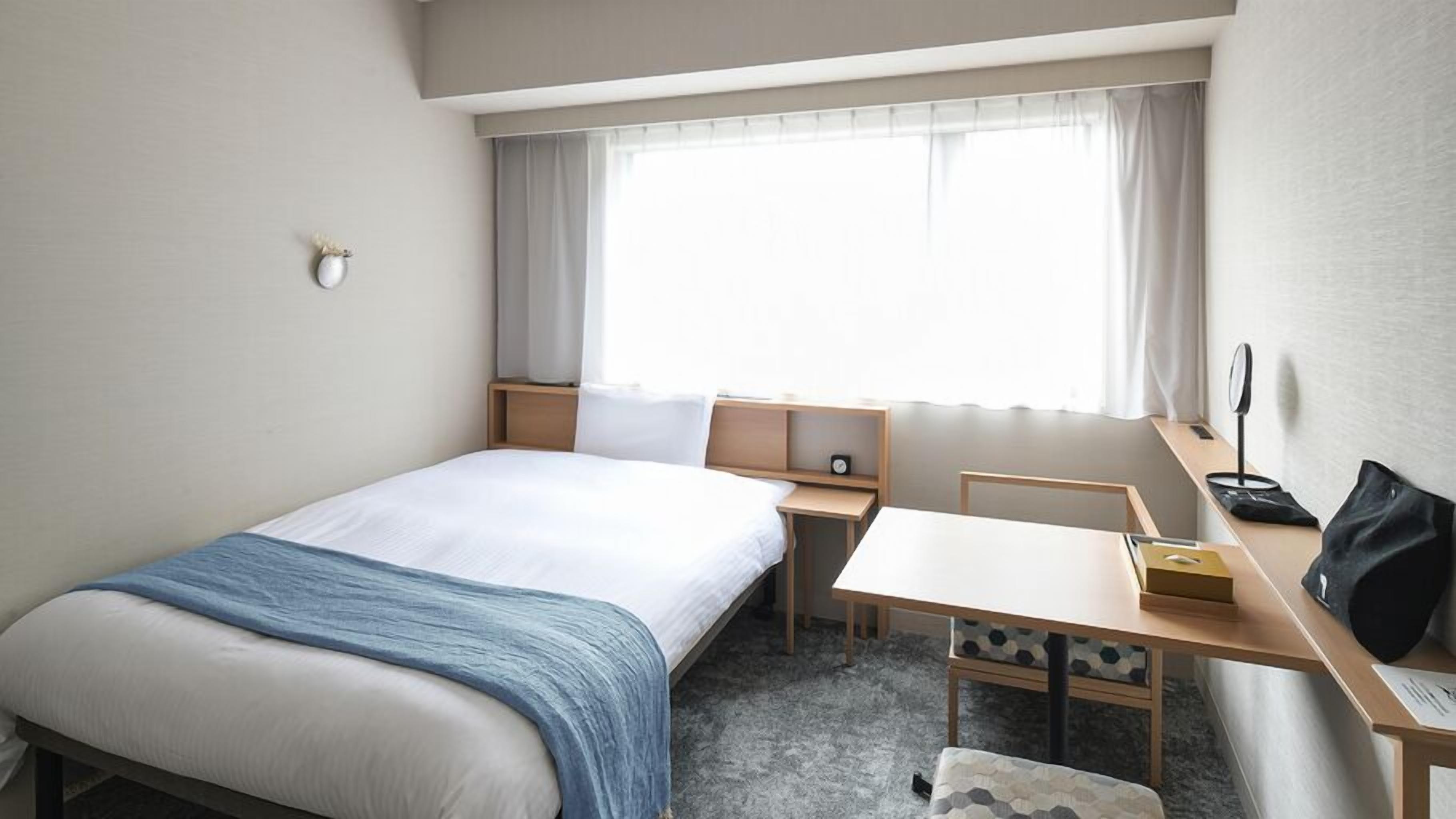 Habitación de hotel con cama, escritorio, silla, mesita, mesita de noche, ventana con cortinas y lámpara de pared.