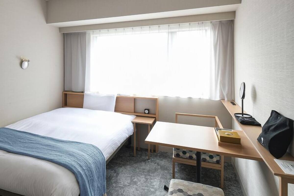 Habitación de hotel con cama, escritorio, silla, mesita, mesita de noche, ventana con cortinas y lámpara de pared.