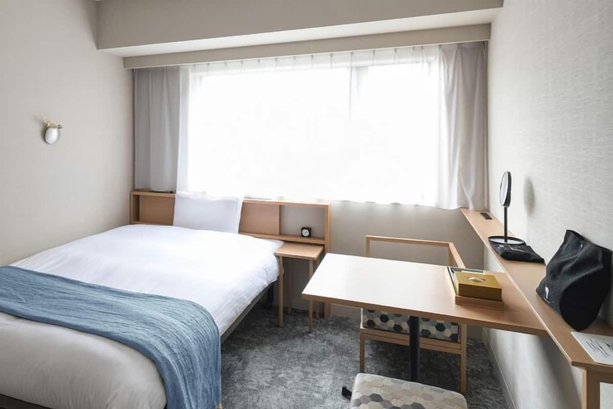 A hotel room with a bed, a desk, a chair, a small table, a nightstand, a window with curtains, and a wall-mounted lamp.