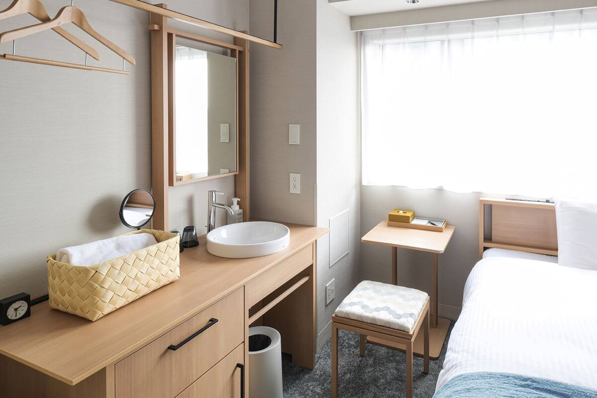 Habitación de hotel con tocador de madera, lavamanos redondo, espejo, mesita de noche con lámpara, un taburete pequeño y cama con ropa blanca.
